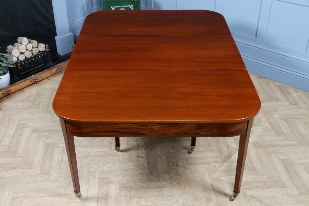 An extended Georgian mahogany dining table featuring semi-circular D-ends, a central drop-leaf section, square tapered legs, and brass castors. View XXIII