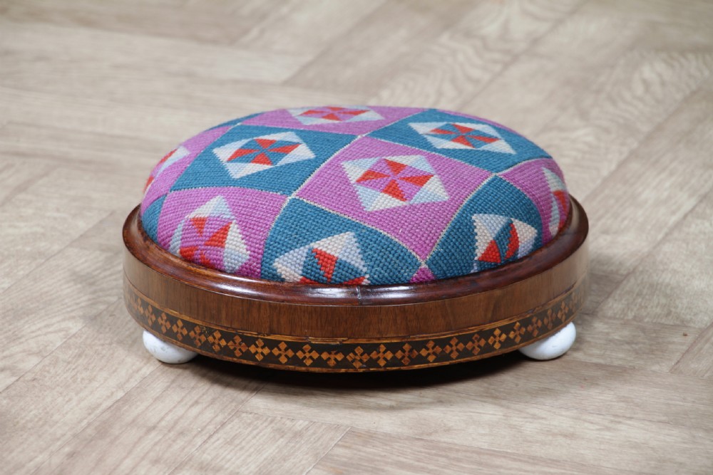 A small round Victorian walnut footstool featuring Tunbridge ware geometric inlay, white ceramic bun feet, and a multi-coloured geometric needlepoint cushion.