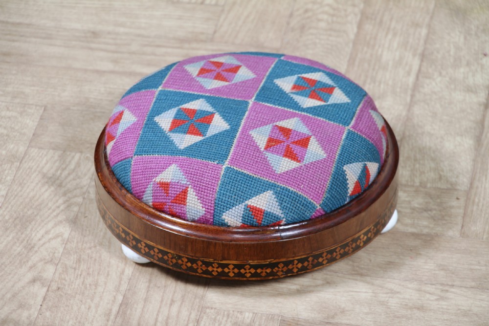A small round Victorian walnut footstool featuring Tunbridge ware geometric inlay, white ceramic bun feet, and a multi-coloured geometric needlepoint cushion. View II