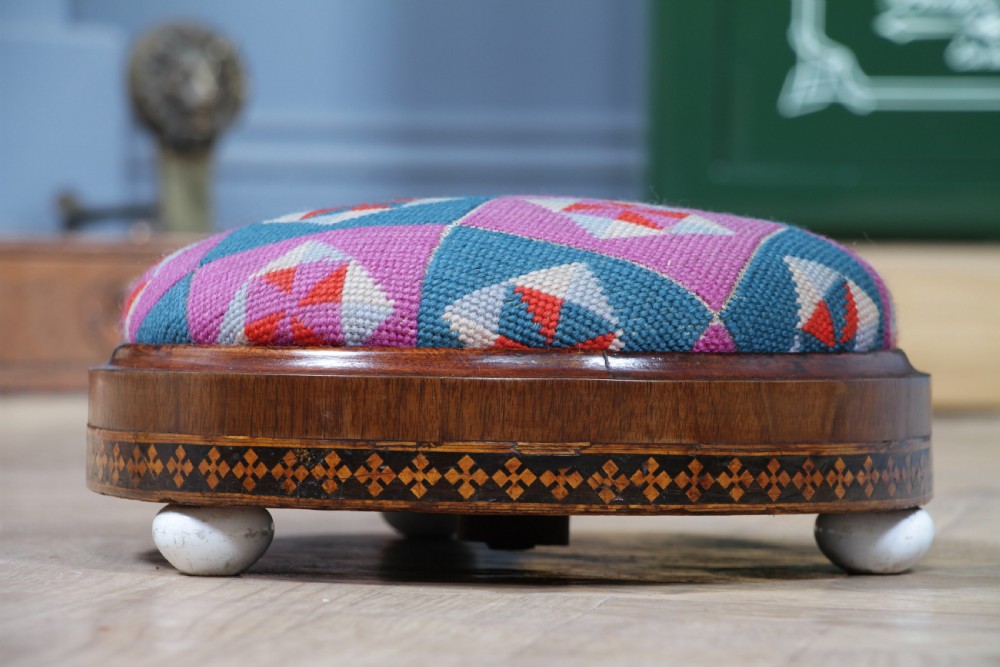 A small round Victorian walnut footstool featuring Tunbridge ware geometric inlay, white ceramic bun feet, and a multi-coloured geometric needlepoint cushion. View III
