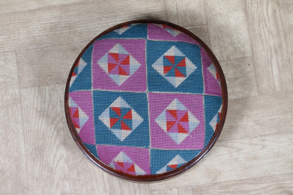 A small round Victorian walnut footstool featuring Tunbridge ware geometric inlay, white ceramic bun feet, and a multi-coloured geometric needlepoint cushion. View V