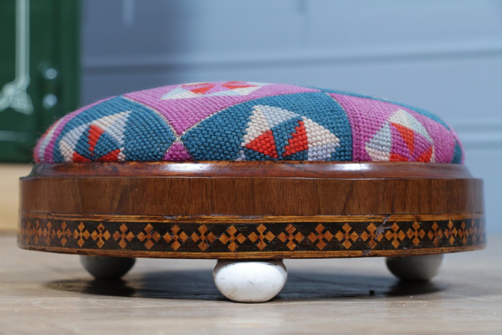 A small round Victorian walnut footstool featuring Tunbridge ware geometric inlay, white ceramic bun feet, and a multi-coloured geometric needlepoint cushion. View VI