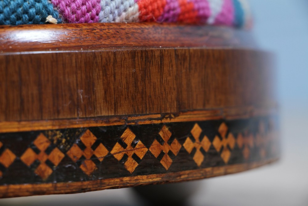 A small round Victorian walnut footstool featuring Tunbridge ware geometric inlay, white ceramic bun feet, and a multi-coloured geometric needlepoint cushion. View VII