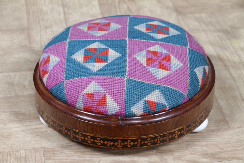 A small round Victorian walnut footstool featuring Tunbridge ware geometric inlay, white ceramic bun feet, and a multi-coloured geometric needlepoint cushion. View XI