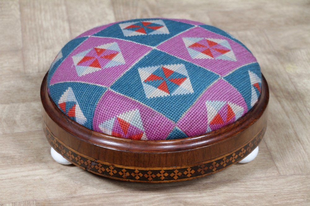 A small round Victorian walnut footstool featuring Tunbridge ware geometric inlay, white ceramic bun feet, and a multi-coloured geometric needlepoint cushion. View XII