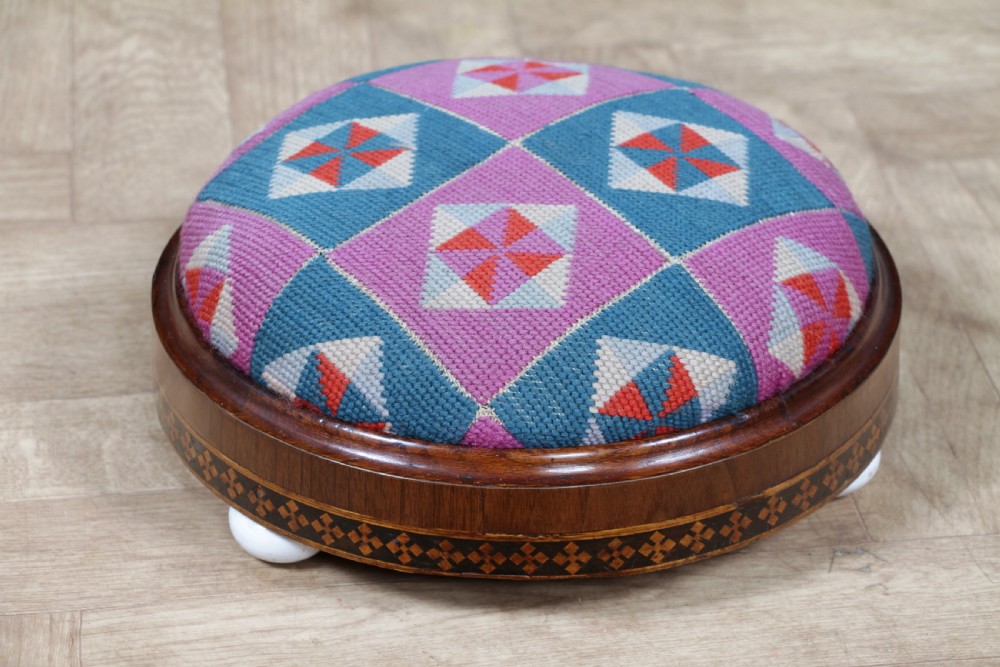 A small round Victorian walnut footstool featuring Tunbridge ware geometric inlay, white ceramic bun feet, and a multi-coloured geometric needlepoint cushion. View XIII