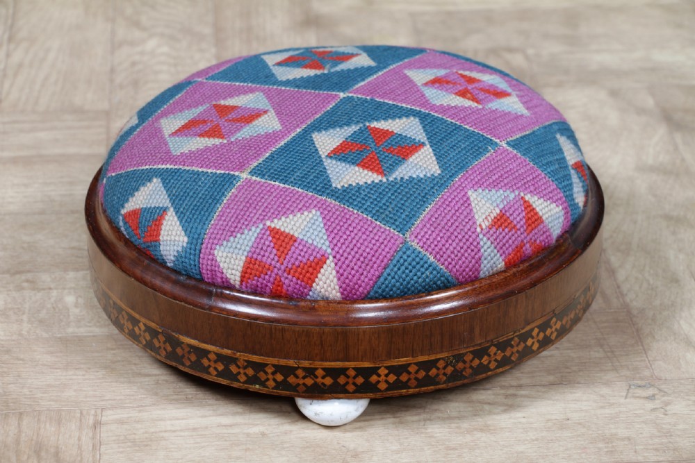 A small round Victorian walnut footstool featuring Tunbridge ware geometric inlay, white ceramic bun feet, and a multi-coloured geometric needlepoint cushion. View XIV