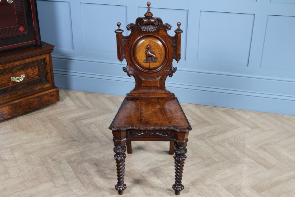 An antique carved dark oak hall chair featuring a decorative backrest with a central crest motif, finials, and barley twist front legs. View VIII