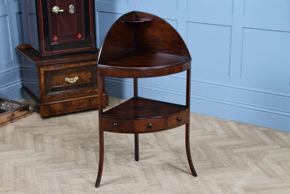 An antique dark mahogany corner washstand featuring a curved splashback with a small top shelf, a middle surface tier, a lower shelf with a central drawer, and splayed legs.