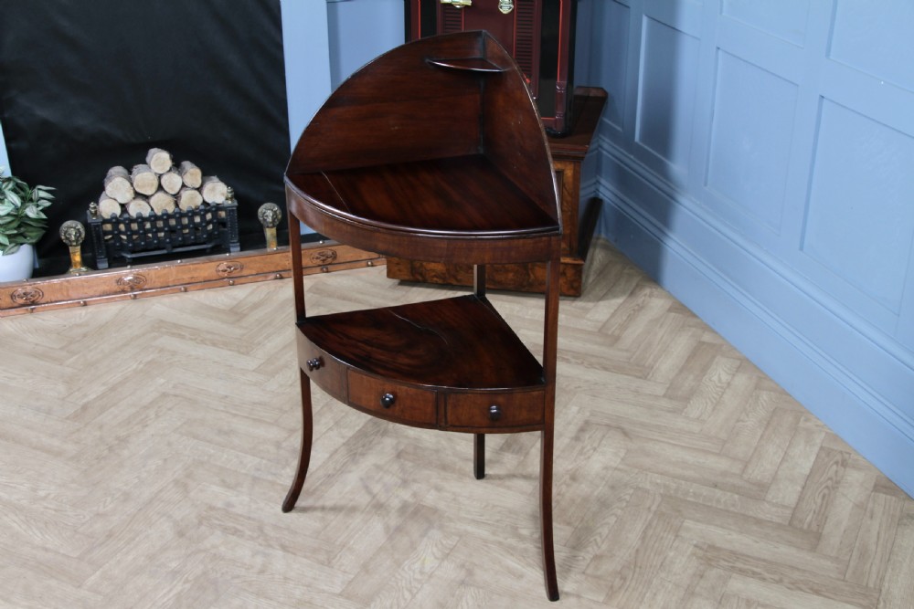 An antique dark mahogany corner washstand featuring a curved splashback with a small top shelf, a middle surface tier, a lower shelf with a central drawer, and splayed legs. View II