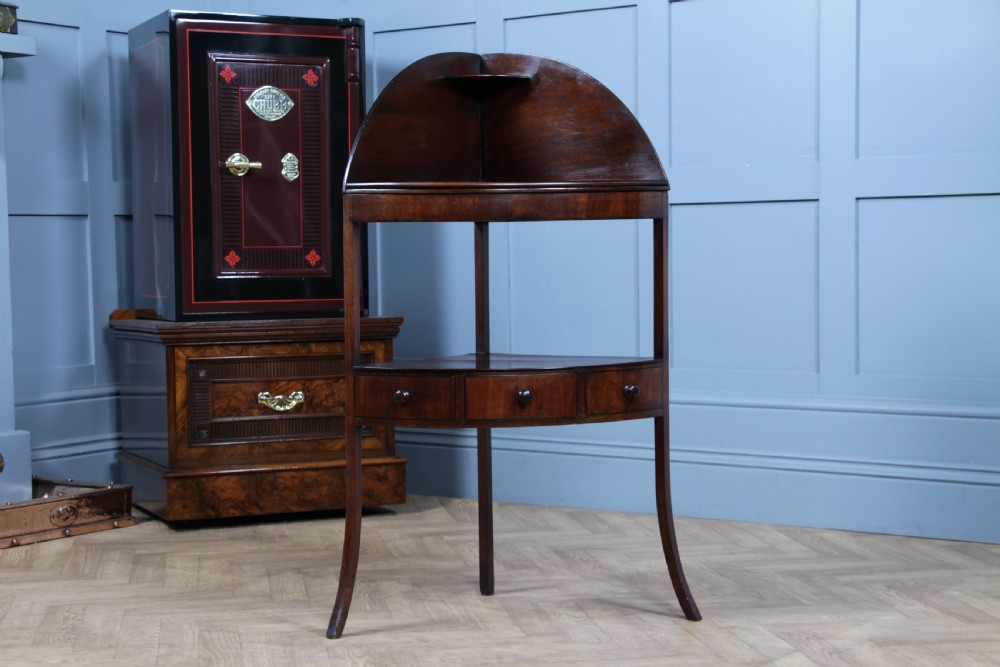 An antique dark mahogany corner washstand featuring a curved splashback with a small top shelf, a middle surface tier, a lower shelf with a central drawer, and splayed legs. View III