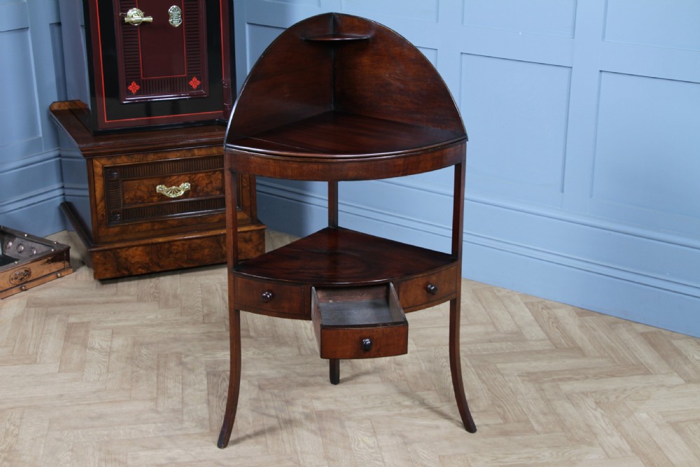 An antique dark mahogany corner washstand featuring a curved splashback with a small top shelf, a middle surface tier, a lower shelf with a central drawer, and splayed legs. View V