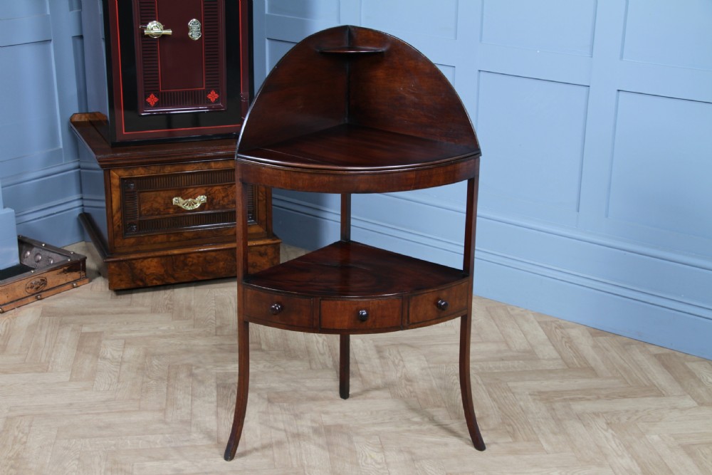 An antique dark mahogany corner washstand featuring a curved splashback with a small top shelf, a middle surface tier, a lower shelf with a central drawer, and splayed legs. View VIII
