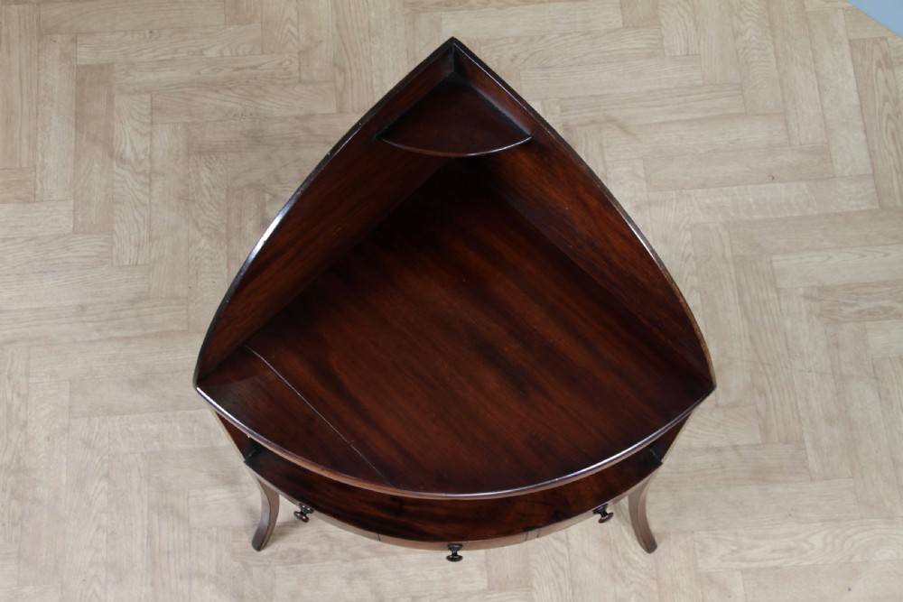 An antique dark mahogany corner washstand featuring a curved splashback with a small top shelf, a middle surface tier, a lower shelf with a central drawer, and splayed legs. View X