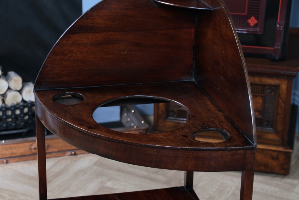 An antique dark mahogany corner washstand featuring a curved splashback with a small top shelf, a middle surface tier, a lower shelf with a central drawer, and splayed legs. View XVI