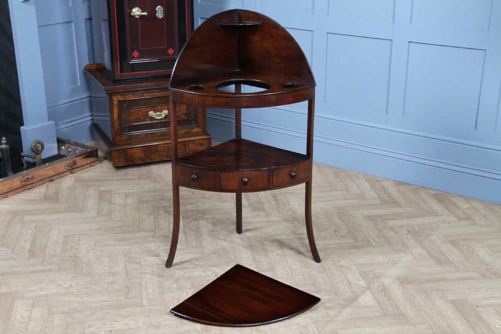 An antique dark mahogany corner washstand featuring a curved splashback with a small top shelf, a middle surface tier, a lower shelf with a central drawer, and splayed legs. View XIX