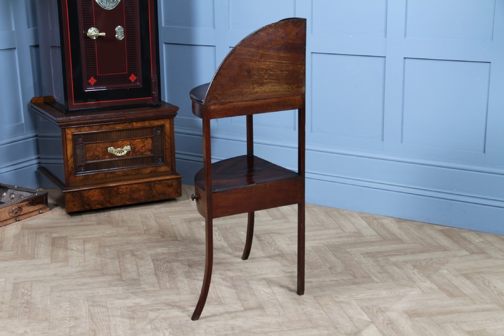 An antique dark mahogany corner washstand featuring a curved splashback with a small top shelf, a middle surface tier, a lower shelf with a central drawer, and splayed legs. View XXI
