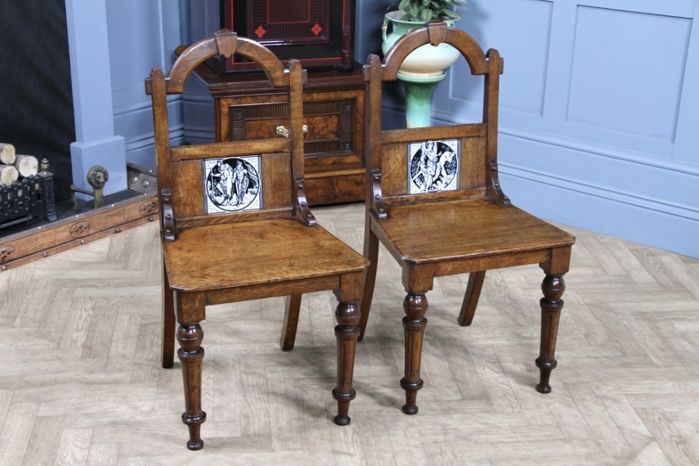 A pair of Victorian Arts and Crafts solid oak hall chairs featuring arched backrests inset with monochrome ceramic Minton tiles and turned front legs.