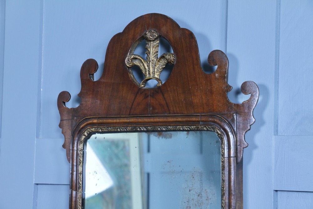 A George III walnut wall mirror featuring silhouette scrollwork, a pierced crest with a gilded plume motif, and a glass plate with visible oxidisation. View VIII