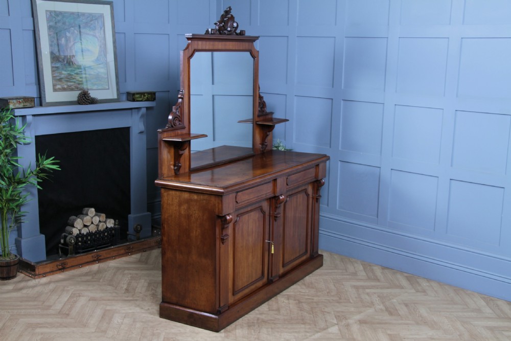 A Victorian mahogany mirror-back sideboard featuring two panelled cabinet doors, upper drawers, tiered side shelves, and a decorative carved cresting. View X