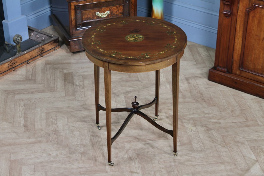 An Edwardian mahogany circular occasional table featuring a painted floral garland rim, central medallion, slender tapered legs, and an X-shaped cross-stretcher with a finial.