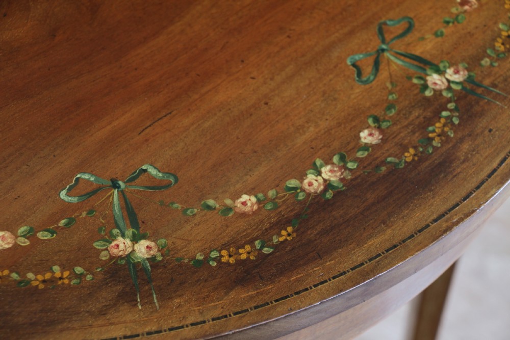 An Edwardian mahogany circular occasional table featuring a painted floral garland rim, central medallion, slender tapered legs, and an X-shaped cross-stretcher with a finial. View V