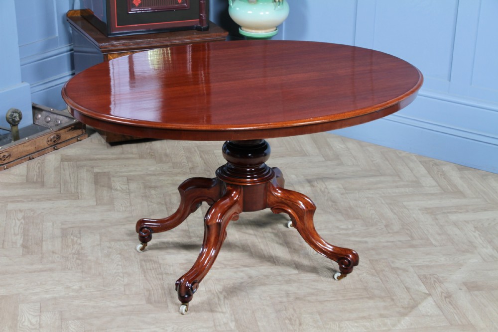 An oval mahogany tilt-top dining table featuring a polished surface, a central turned pedestal, and four carved cabriole legs on white ceramic castors.