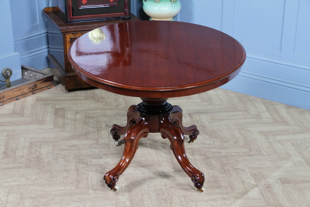 An oval mahogany tilt-top dining table featuring a polished surface, a central turned pedestal, and four carved cabriole legs on white ceramic castors. View II