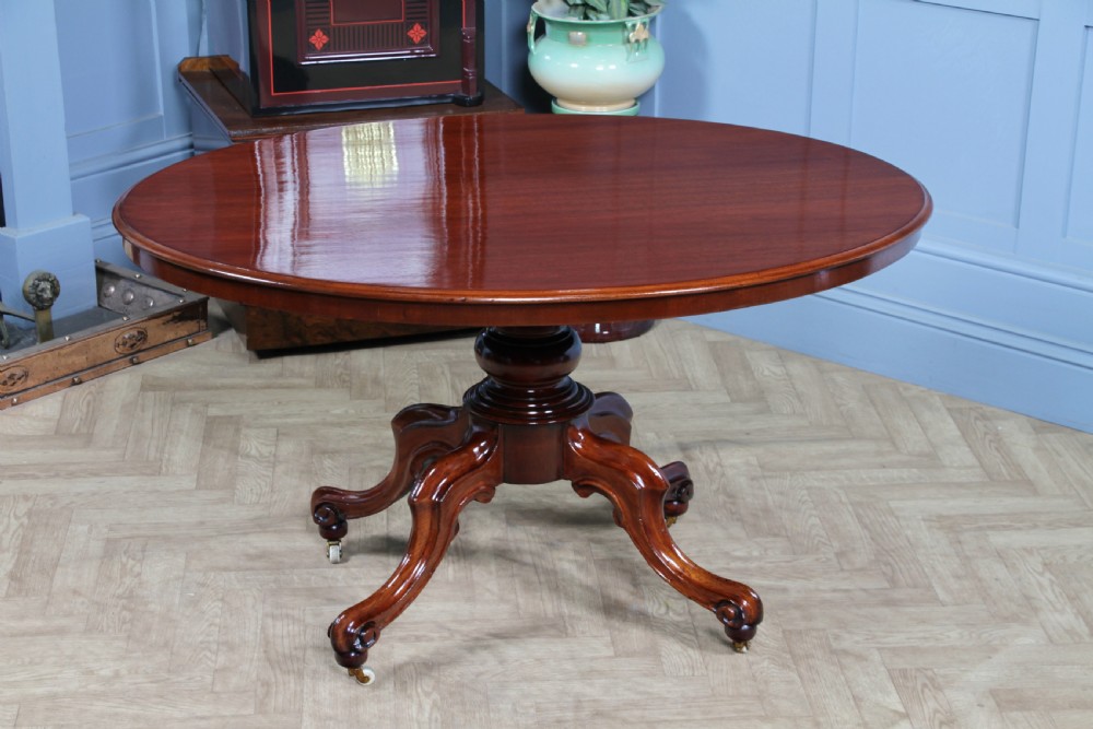An oval mahogany tilt-top dining table featuring a polished surface, a central turned pedestal, and four carved cabriole legs on white ceramic castors. View III