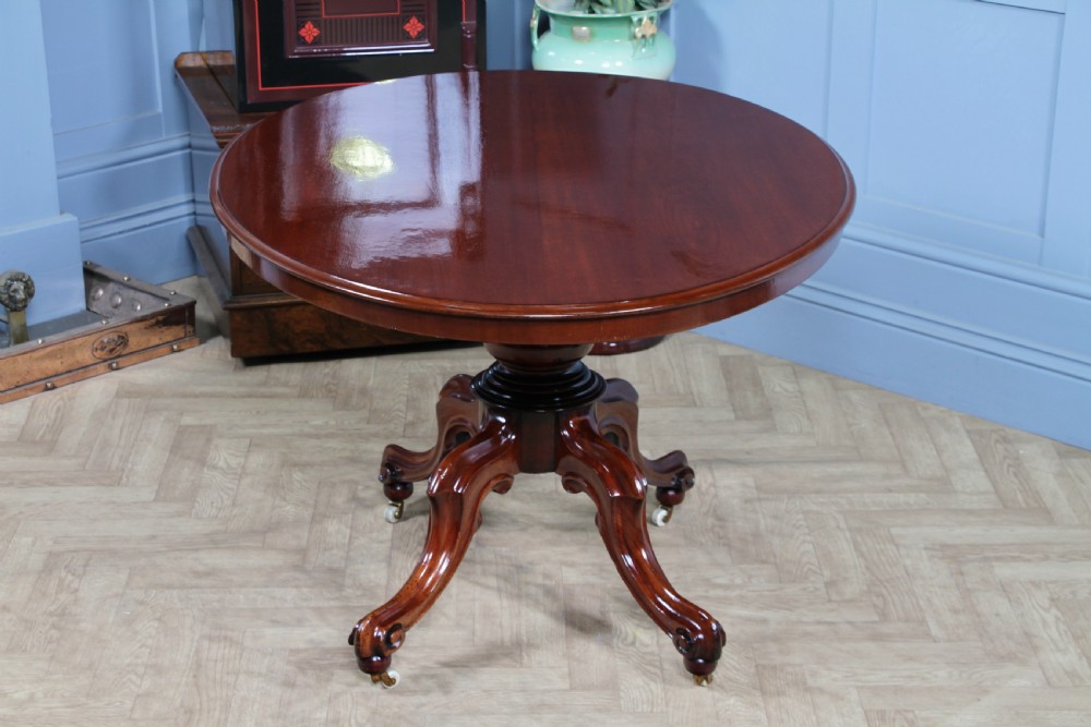 An oval mahogany tilt-top dining table featuring a polished surface, a central turned pedestal, and four carved cabriole legs on white ceramic castors. View V