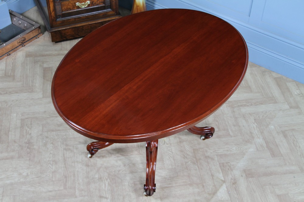 An oval mahogany tilt-top dining table featuring a polished surface, a central turned pedestal, and four carved cabriole legs on white ceramic castors. View VI
