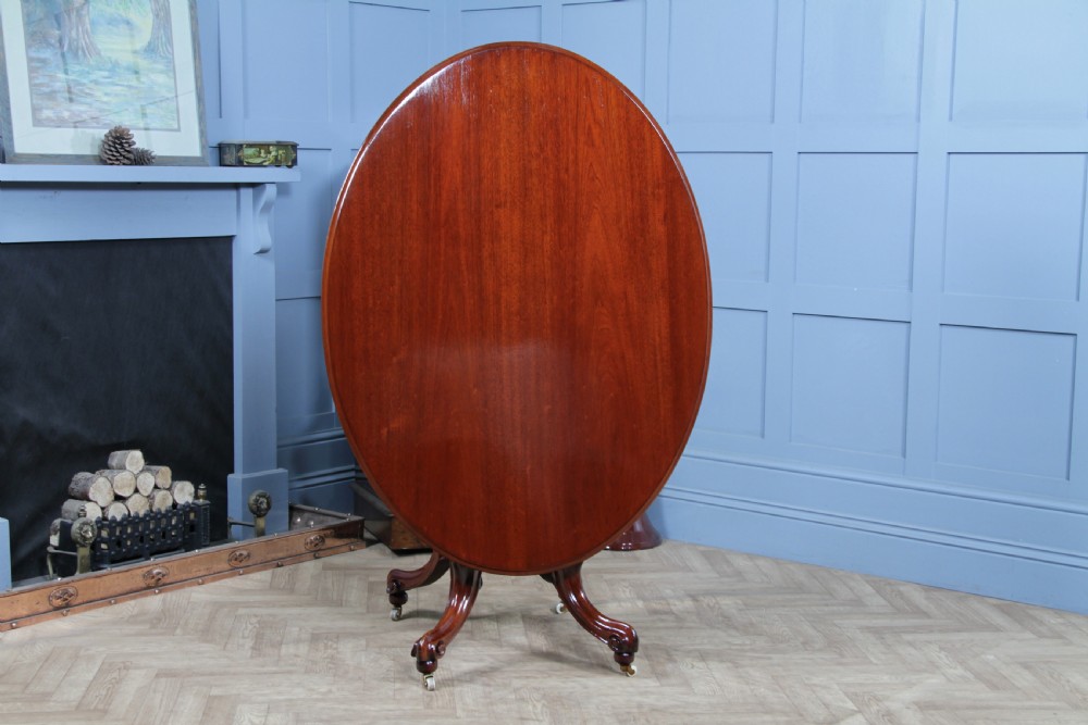 An oval mahogany tilt-top dining table featuring a polished surface, a central turned pedestal, and four carved cabriole legs on white ceramic castors. View VII