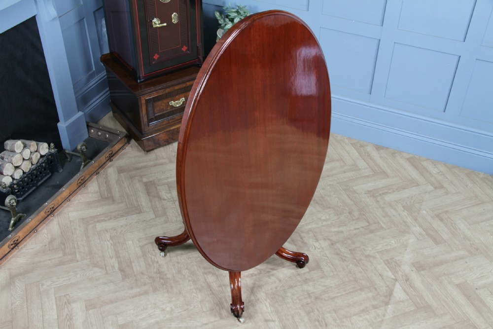 An oval mahogany tilt-top dining table featuring a polished surface, a central turned pedestal, and four carved cabriole legs on white ceramic castors. View VIII