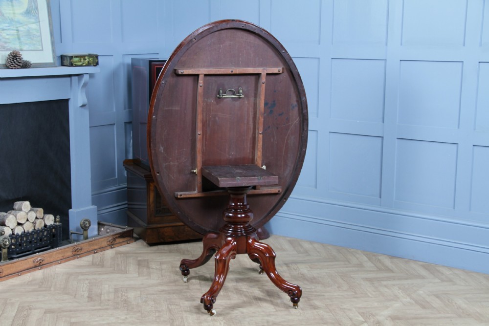An oval mahogany tilt-top dining table featuring a polished surface, a central turned pedestal, and four carved cabriole legs on white ceramic castors. View IX