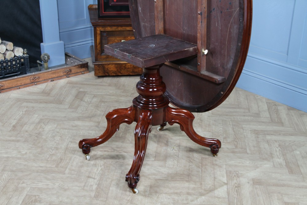 An oval mahogany tilt-top dining table featuring a polished surface, a central turned pedestal, and four carved cabriole legs on white ceramic castors. View X