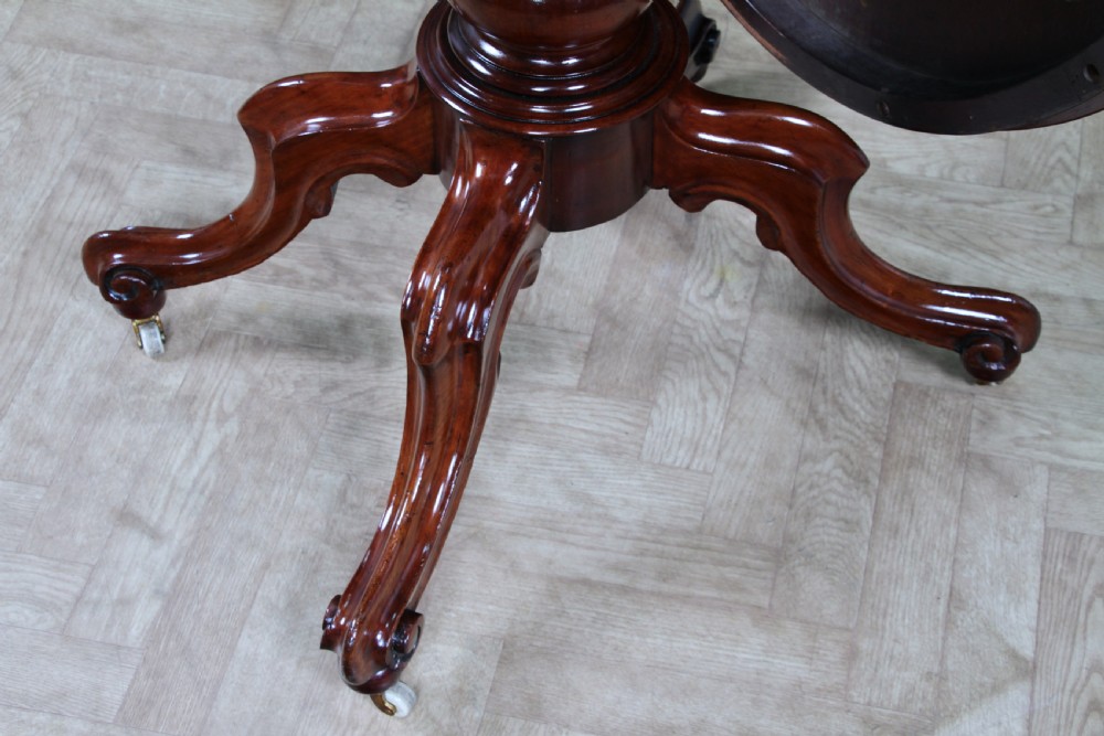 An oval mahogany tilt-top dining table featuring a polished surface, a central turned pedestal, and four carved cabriole legs on white ceramic castors. View XI