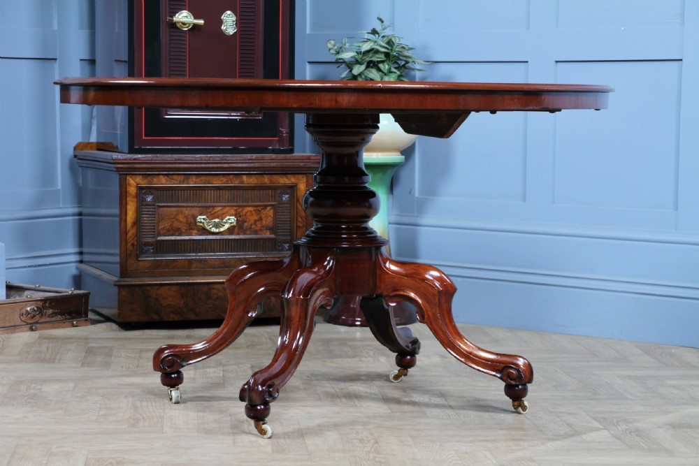 An oval mahogany tilt-top dining table featuring a polished surface, a central turned pedestal, and four carved cabriole legs on white ceramic castors. View XII