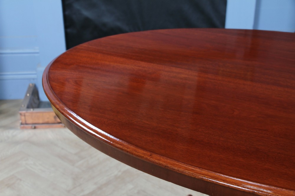An oval mahogany tilt-top dining table featuring a polished surface, a central turned pedestal, and four carved cabriole legs on white ceramic castors. View XV