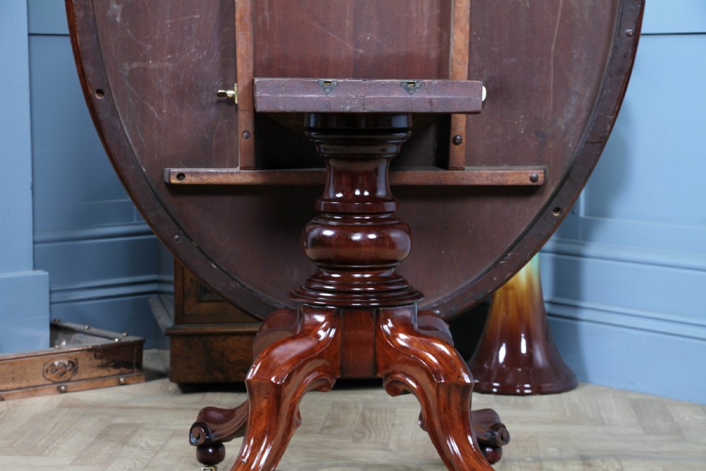 An oval mahogany tilt-top dining table featuring a polished surface, a central turned pedestal, and four carved cabriole legs on white ceramic castors. View XVI