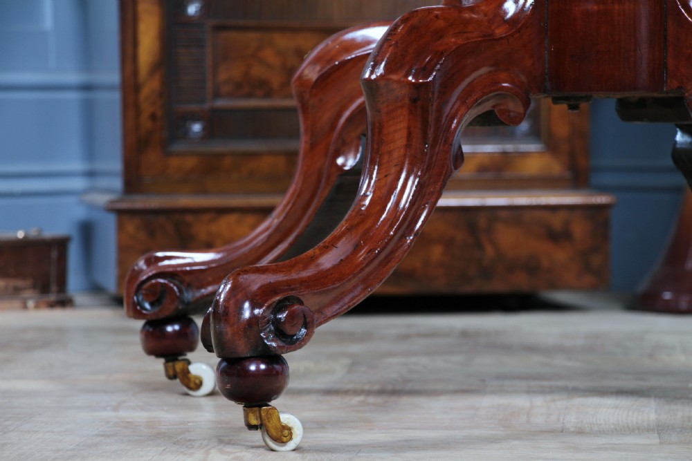 An oval mahogany tilt-top dining table featuring a polished surface, a central turned pedestal, and four carved cabriole legs on white ceramic castors. View XVIII