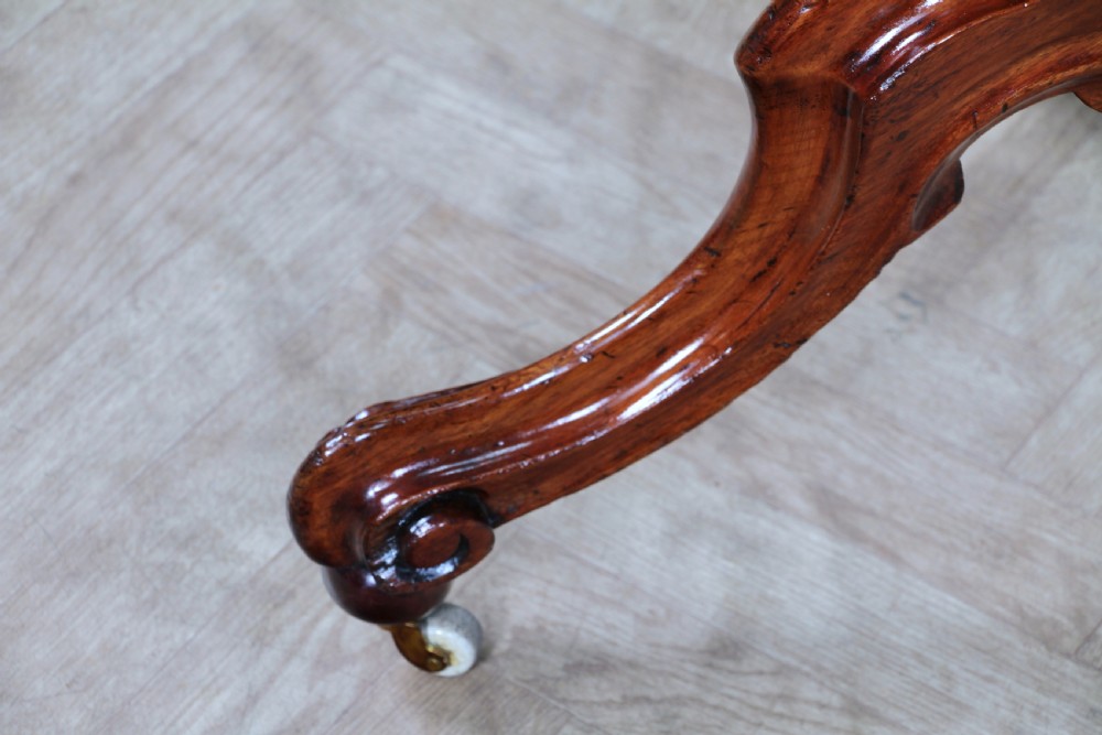 An oval mahogany tilt-top dining table featuring a polished surface, a central turned pedestal, and four carved cabriole legs on white ceramic castors. View XIX