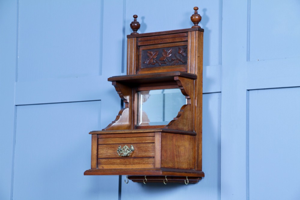 An Edwardian wall-mounted wooden cabinet featuring top finials, a carved floral panel, a central mirror, a lower drawer box with a brass handle, and bottom key hooks.