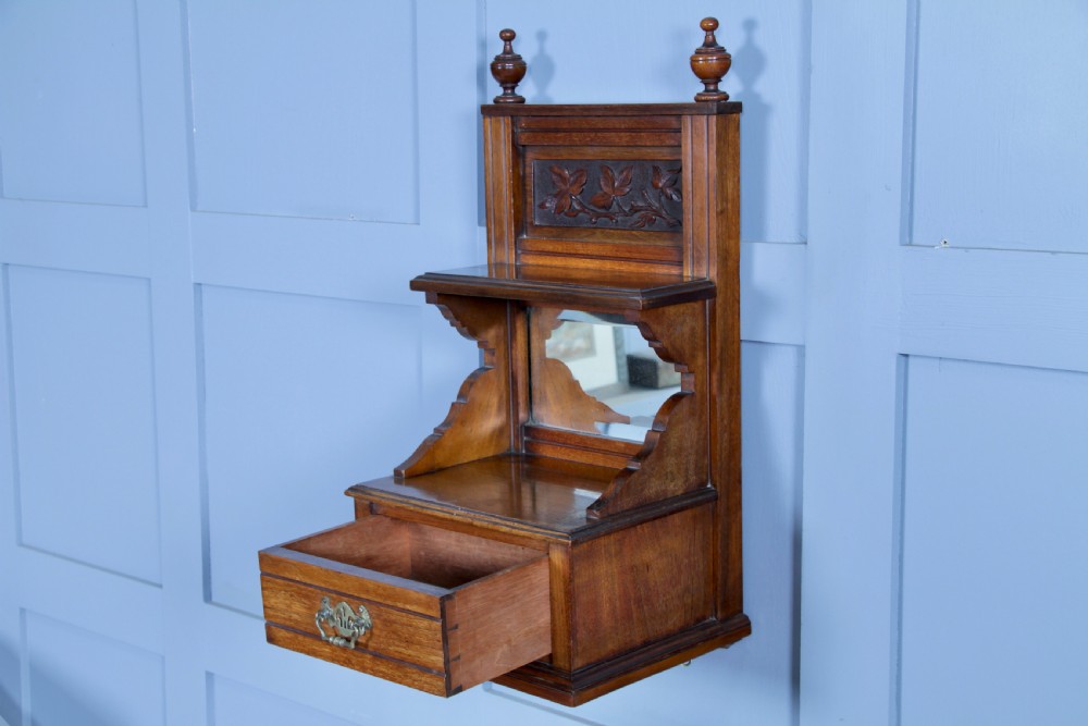 An Edwardian wall-mounted wooden cabinet featuring top finials, a carved floral panel, a central mirror, a lower drawer box with a brass handle, and bottom key hooks. View II