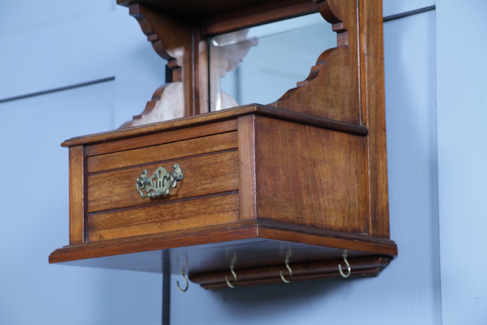 An Edwardian wall-mounted wooden cabinet featuring top finials, a carved floral panel, a central mirror, a lower drawer box with a brass handle, and bottom key hooks. View V