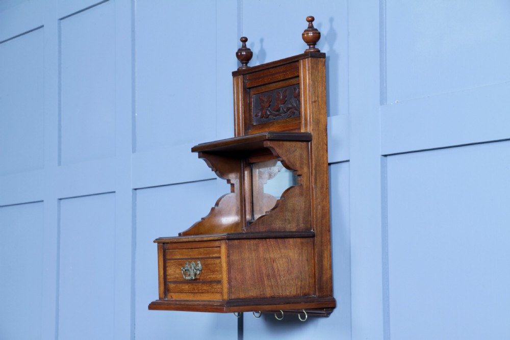 An Edwardian wall-mounted wooden cabinet featuring top finials, a carved floral panel, a central mirror, a lower drawer box with a brass handle, and bottom key hooks. View VIII