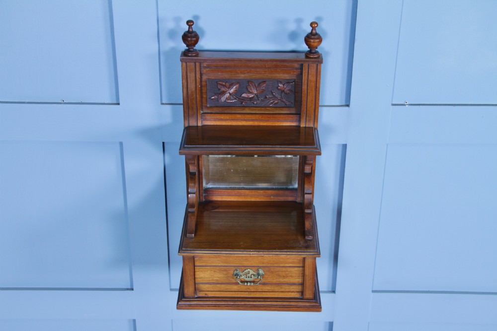 An Edwardian wall-mounted wooden cabinet featuring top finials, a carved floral panel, a central mirror, a lower drawer box with a brass handle, and bottom key hooks. View IX