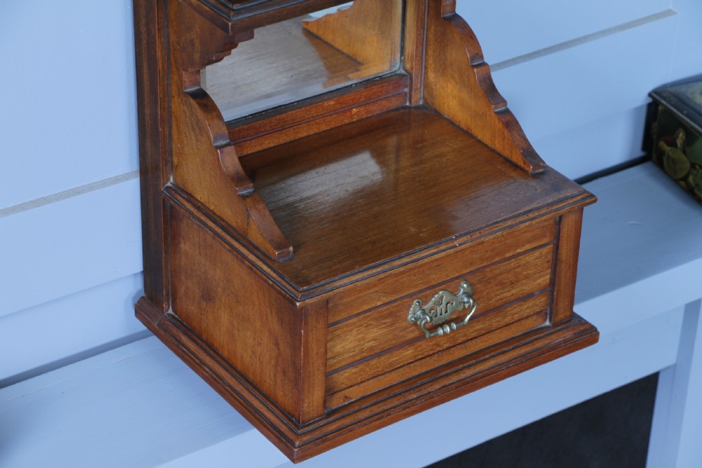 An Edwardian wall-mounted wooden cabinet featuring top finials, a carved floral panel, a central mirror, a lower drawer box with a brass handle, and bottom key hooks. View XIII
