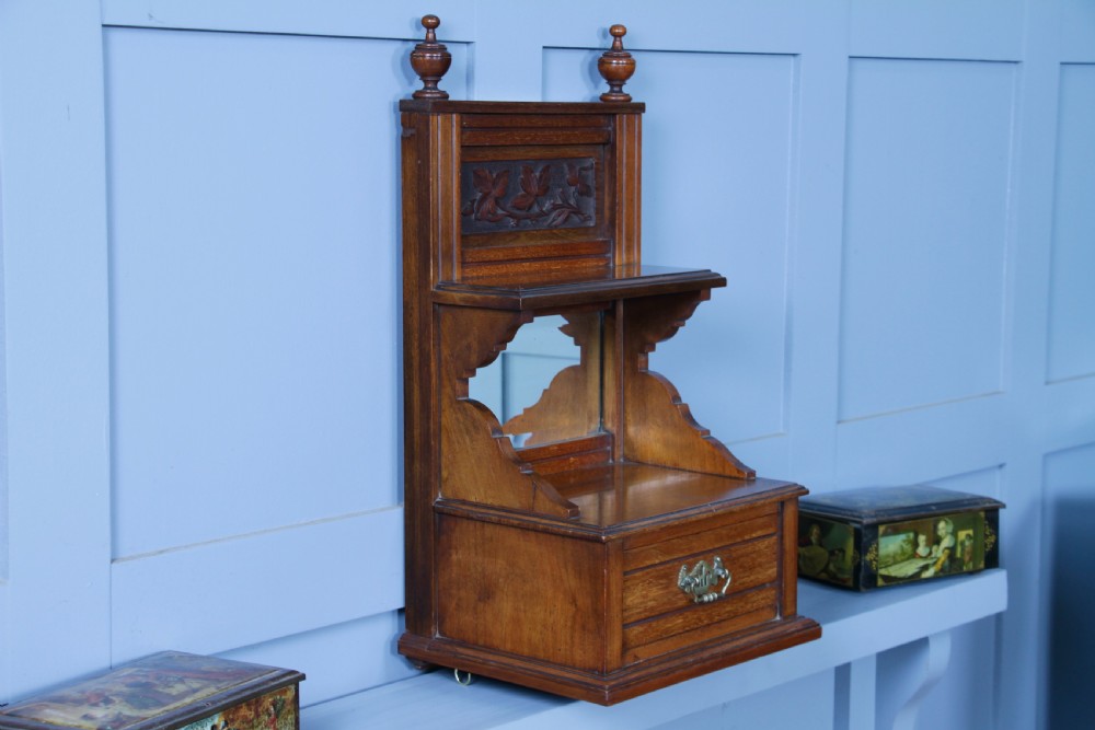 An Edwardian wall-mounted wooden cabinet featuring top finials, a carved floral panel, a central mirror, a lower drawer box with a brass handle, and bottom key hooks. View XX