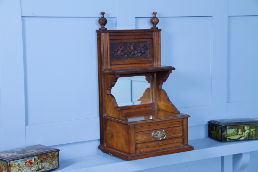 An Edwardian wall-mounted wooden cabinet featuring top finials, a carved floral panel, a central mirror, a lower drawer box with a brass handle, and bottom key hooks. View XXI