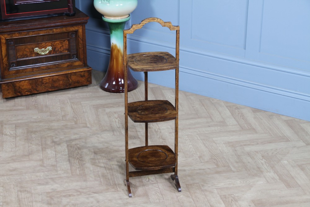 An Edwardian solid oak three-tier folding cake stand featuring square tiered shelves with recessed circular plate holders and an arched top handle. View II